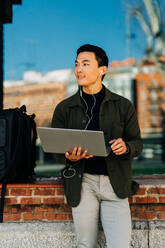 Nachdenklicher junger asiatischer männlicher Freiberufler in Freizeitkleidung, der wegschaut, während er aus der Ferne am Laptop arbeitet, der in der Nähe eines Gebäudes mit Glasspiegel steht - ADSF26027