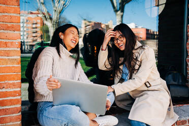 Fröhliche junge asiatische beste Freundinnen mit langen dunklen Haaren in stilvollen Outfits lachen, während sie ein lustiges Video auf einem Laptop ansehen, der in der Nähe eines Backsteingebäudes auf einer Straße in der Stadt steht - ADSF26031