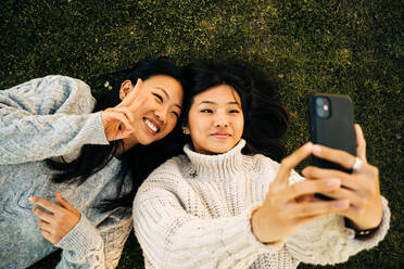 Von oben von glücklichen jungen asiatischen Damen in stilvollen gestrickten Pullover lächelnd und zeigt v Zeichen Geste bei der Aufnahme von Selfie auf Smartphone auf Gras im Park liegen - ADSF26033