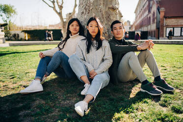 Ganzer Körper von ruhigen jungen asiatischen weiblichen und männlichen Studenten in trendigen Outfits entspannen auf grasbewachsenen Rasen in der Nähe von Baum, während die Zeit zusammen auf dem Campus an einem sonnigen Tag - ADSF26034