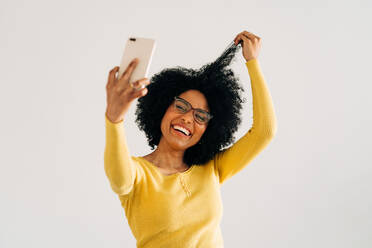 Positive afroamerikanische Frau, die ein Smartphone benutzt, während sie ein Selfie mit einem Mobiltelefon auf weißem Hintergrund macht - ADSF26037