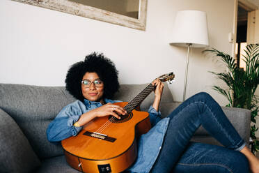 Delightful African American female composing song on guitar on sofa in living room - ADSF26038