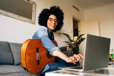 Smiling African American female musician with short curly hair in eyeglasses and casual clothes sitting on comfortable sofa while singing song and playing guitar in front of laptop - ADSF26039