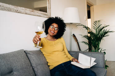 Fröhliche afroamerikanische Frau in leuchtend gelbem Pullover und Brille auf weichem grauen Sofa im Wohnzimmer - ADSF26040