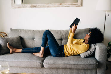 Seitenansicht einer schlanken Afroamerikanerin, die sich auf einem weichen grauen Sofa ausruht und Tagebuch liest - ADSF26041