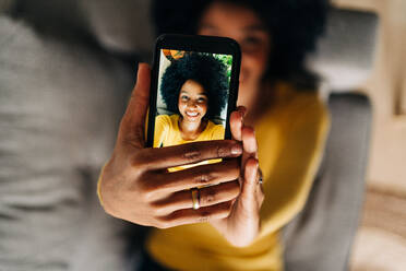 Draufsicht auf eine glückliche afroamerikanische Frau, die auf dem Sofa liegend ein Selbstporträt mit ihrem Mobiltelefon aufnimmt - ADSF26042