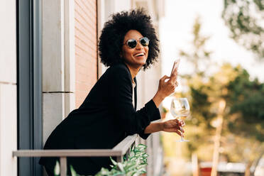 Happy African American female with glass of wine in elegant suit browsing on mobile phone on balcony - ADSF26045