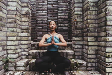 Young woman meditating with hands clasped in stone structure - DSIF00494