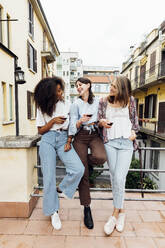 Smiling friends with wineglass leaning on terrace railing - MEUF03446