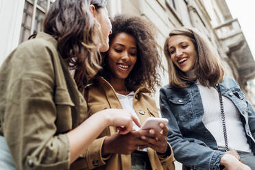 Freunde, die ein Mobiltelefon benutzen, während sie vor einem Gebäude stehen - MEUF03489