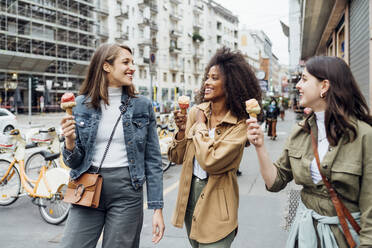 Lächelnde Freunde essen Eis beim Spaziergang in der Stadt Straße - MEUF03506