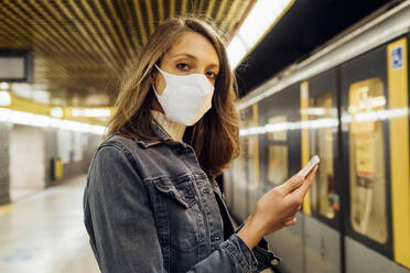 Frau mit Gesichtsmaske hält Mobiltelefon in der U-Bahn - MEUF03522