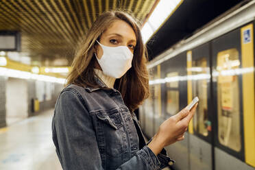 Woman with face mask holding mobile phone while standing in subway - MEUF03522