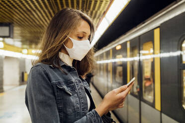 Woman with face mask using mobile phone while standing in subway - MEUF03523