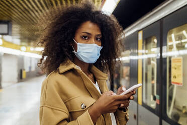 Junge Frau mit Schutzmaske, die in der U-Bahn steht und ein Handy in der Hand hält - MEUF03524