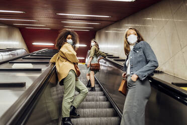 Freunde mit Gesichtsschutz, die in sozialer Distanz auf der Rolltreppe stehen - MEUF03527