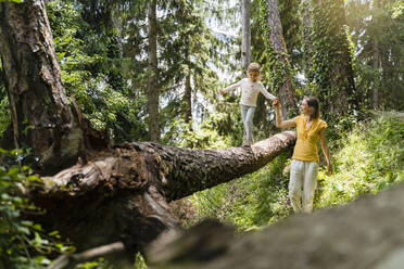 Mother helping daughter walking on fallen tree in forest - DIGF16069