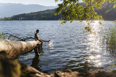 Man dangling legs and splashing water while sitting on fallen tree at lakeshore - DIGF16088