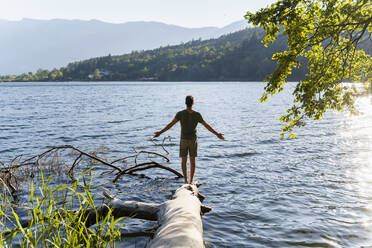 Carefree man with arms outstretched standing on fallen tree at lakeshore - DIGF16089