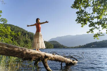 Carefree woman with arms outstretched standing on fallen tree at lake - DIGF16095