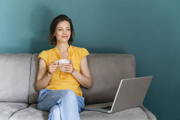 Woman holding coffee cup while sitting by laptop on sofa - DIGF16101