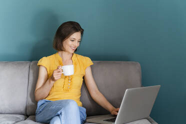 Woman with coffee cup using laptop while sitting on sofa - DIGF16102