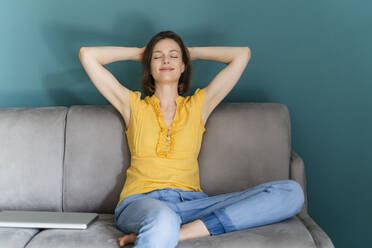 Woman with hands behind head sitting by laptop on sofa at home - DIGF16108