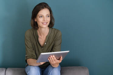 Thoughtful woman holding digital tablet while sitting in front of blue wall - DIGF16122