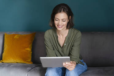 Smiling woman using digital tablet while sitting on sofa in living room - DIGF16125