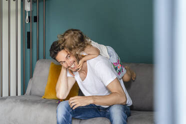 Father piggybacking playful daughter while sitting on sofa in living room - DIGF16127