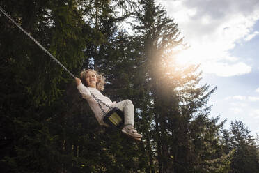 Smiling girl swinging on rope swing in forest - DIGF16142