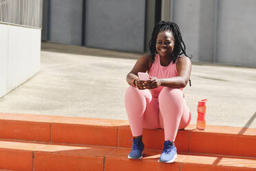 Lächelnde junge Frau in Sportkleidung, die ihr Handy hält, während sie auf einem Fußweg sitzt - AODF00539