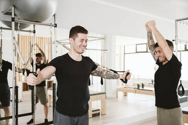 Lächelnder männlicher Ausbilder, der einem Mann beim Training im Pilates-Studio hilft - EBBF04292