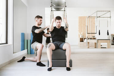 Männlicher Fitnesstrainer, der einem Mann bei der Ausübung von Pilates im Studio hilft - EBBF04301