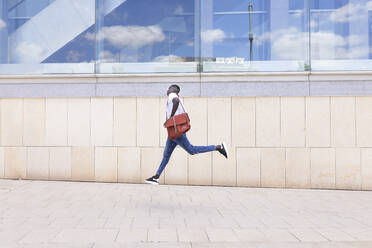 Young man with shoulder bag running by wall - JPTF00883