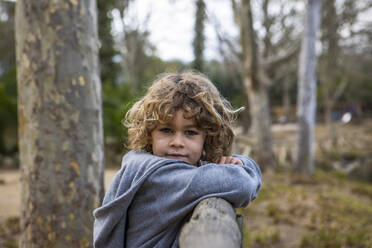 Cute child in casual outfit on old wooden fence looking at camera near trees in the park - ADSF26062