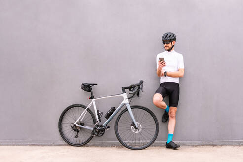 Lächelnder männlicher Radfahrer in Sportkleidung und mit Sonnenbrille, der mit einem Handy und angehobenem Bein am Fahrrad steht, während er im Freien wegschaut - ADSF26070