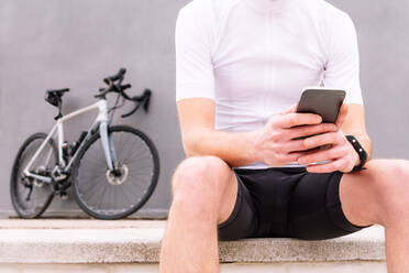 Crop Biker in Sportkleidung sitzt auf dem Bürgersteig und blättert Handy gegen Fahrrad - ADSF26071