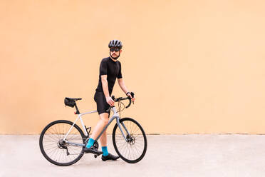 Männlicher Radfahrer in Sportkleidung mit Fahrrad, der auf einem Gehweg in der Stadt spazieren geht, während er auf orangefarbenem Hintergrund wegschaut - ADSF26075