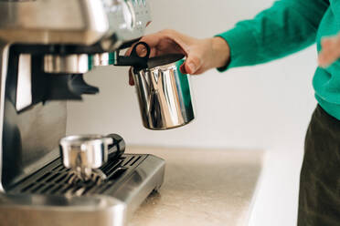 Crop unrecognizable person with metal jug preparing coffee with professional machine at table in house - ADSF26099