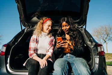 Niedriger Winkel von fröhlichen multirassischen Freundinnen, die im Kofferraum eines Autos sitzen und gemeinsam in sozialen Medien auf ihrem Handy surfen - ADSF26105