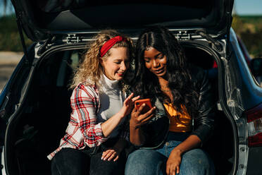 Glückliche multirassische Freundinnen, die im Kofferraum eines Autos sitzen und gemeinsam in sozialen Medien auf ihrem Handy surfen - ADSF26106