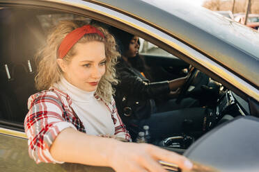 Stilvolle multiethnische Freundinnen sitzen in einem modernen Auto in der Stadt und schauen in den Seitenspiegel - ADSF26107