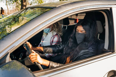Schwarze Fahrerin mit Maske sitzt in einem modernen Auto mit einem Freund in der Stadt und schaut in den Seitenspiegel - ADSF26109