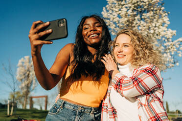 Low angle of cheerful multiracial female best friends embracing in spring garden and taking self shot on smartphone on sunny day - ADSF26118