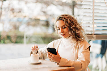 Content female sitting at table in coffee house and stirring tasty drink in cup while browsing on smartphone - ADSF26121