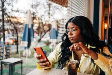 Stilvolle afroamerikanische Frau sitzt an einem Tisch in einem Café mit einem erfrischenden Soda-Getränk und surft in den sozialen Medien auf ihrem Mobiltelefon - ADSF26122