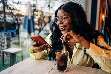 Stilvolle afroamerikanische Frau sitzt an einem Tisch in einem Café mit einem erfrischenden Soda-Getränk und surft in den sozialen Medien auf ihrem Mobiltelefon - ADSF26123