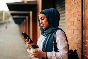 Seitenansicht einer ethnischen Frau im Hidschab, die auf dem Bahnsteig eines Bahnhofs steht und mit ihrem Handy telefoniert, während sie auf den Zug wartet - ADSF26129