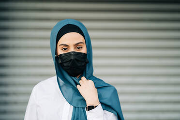 Muslim female wearing protective mask and traditional headscarf standing against wall in city and looking at camera - ADSF26133